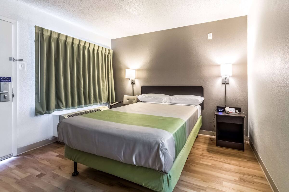Ein Hotelzimmer mit einem Bett, einem Nachttisch, einer Lampe und einem Fenster mit Vorhängen.