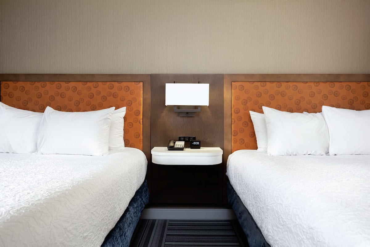 A hotel room with two beds, a small bedside table with a clock and phone, and a wall-mounted TV.
