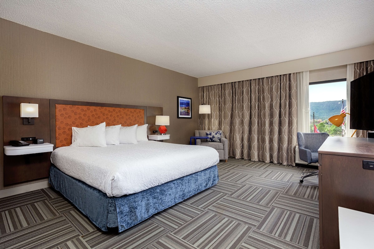 A hotel room with a large bed, a desk, a chair, a TV, and a window with curtains.