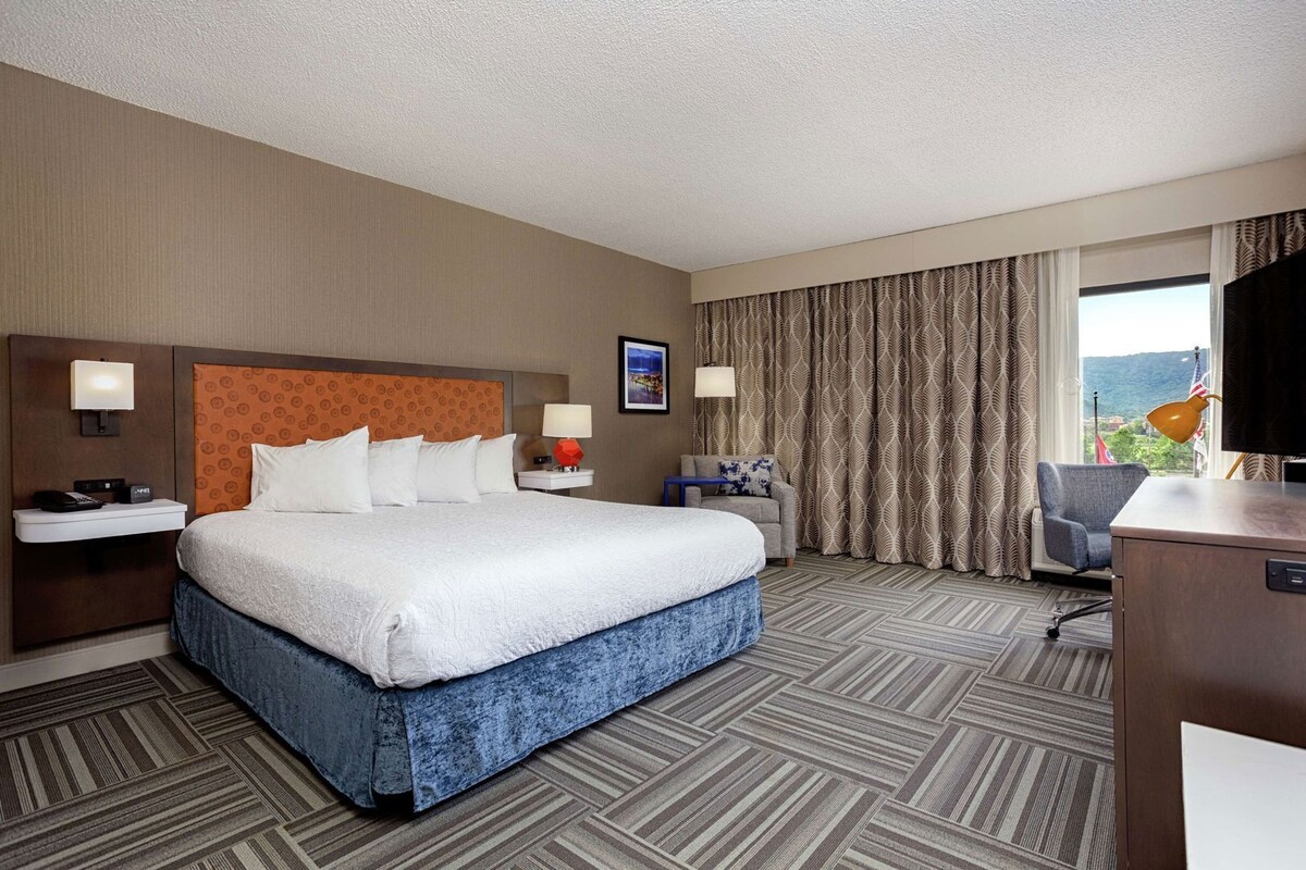 A hotel room with a large bed, a desk, a chair, a TV, and a window with curtains.