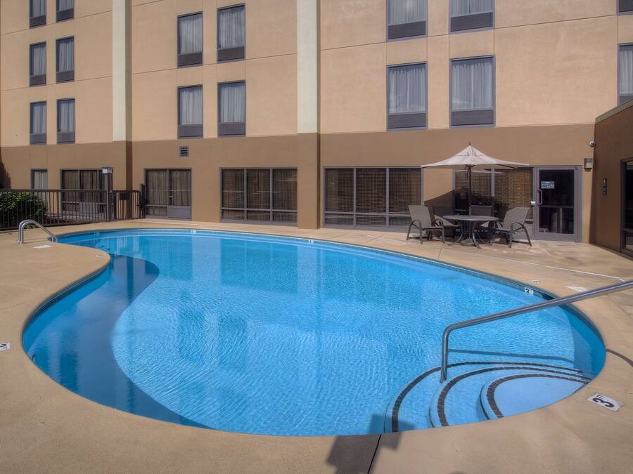Una piscina con borde curvo, rodeada por un edificio color crema con varias ventanas y una zona de estar exterior cubierta.
