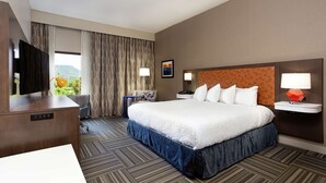 A hotel room with a large bed, a desk with a flat-screen TV, a desk lamp, a nightstand with a lamp, a window with curtains, and a painting on the wall.