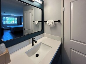 A bathroom with a white sink, a black faucet, and a mirror reflecting a room with a bed and a window.