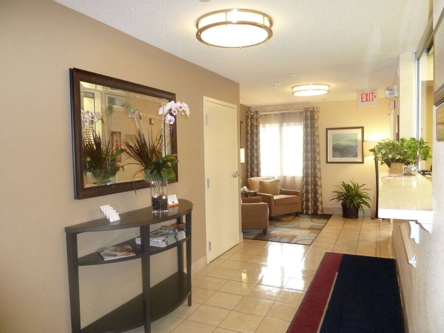 A hotel lobby with a reception desk, a mirror, a vase with flowers, a chair, a couch, and a door with an exit sign.