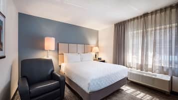 A hotel room with a bed, a chair, two bedside lamps, a window with curtains, and a wall-mounted heater.