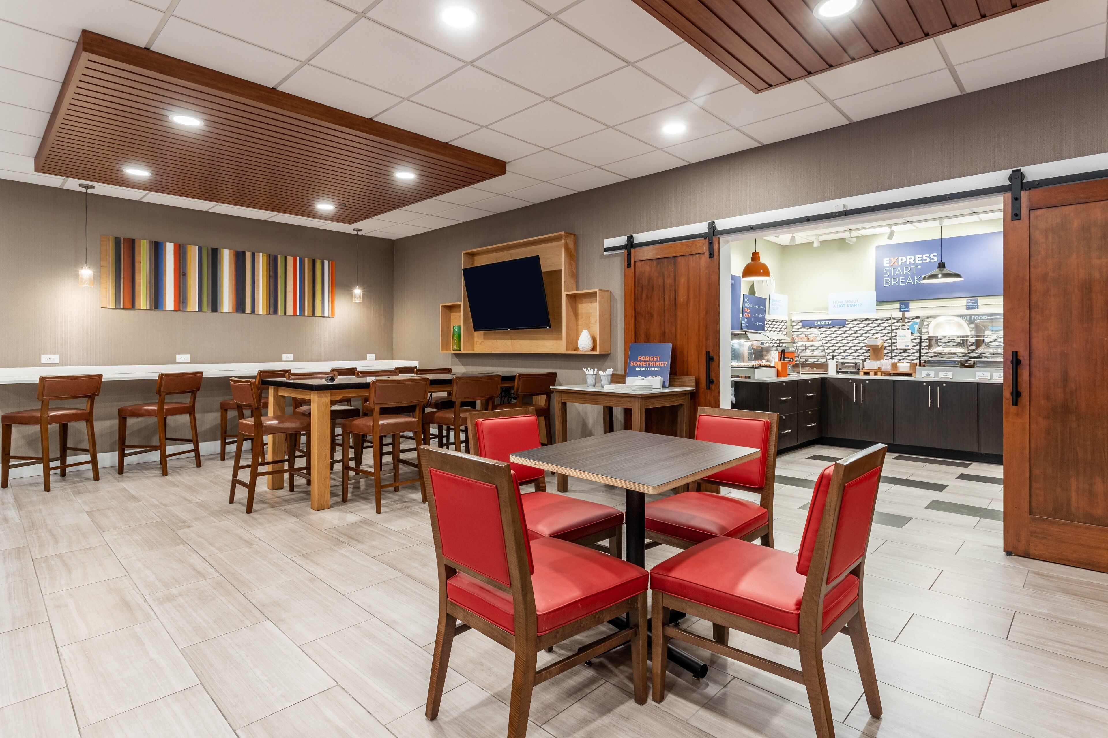 Un restaurante moderno con mesas de madera y sillas rojas, una pantalla de televisión grande y un área de cocina visible a través de una puerta corrediza.