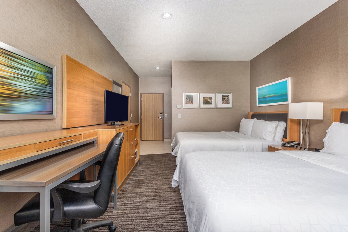 A hotel room with a desk, two beds, a TV, and artwork on the walls.
