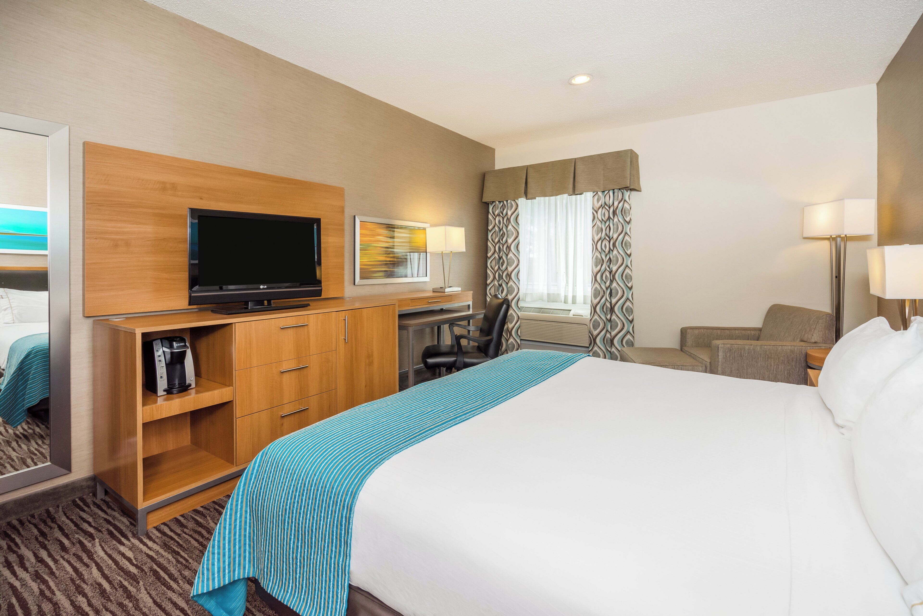 A hotel room with a bed, a desk with a computer, a television, a coffee maker, and a window with curtains.