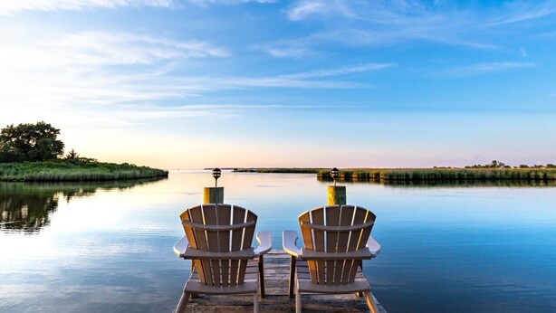 Zwei Adirondack-Stühle, die auf ein ruhiges Gewässer mit blauem Himmel blicken.