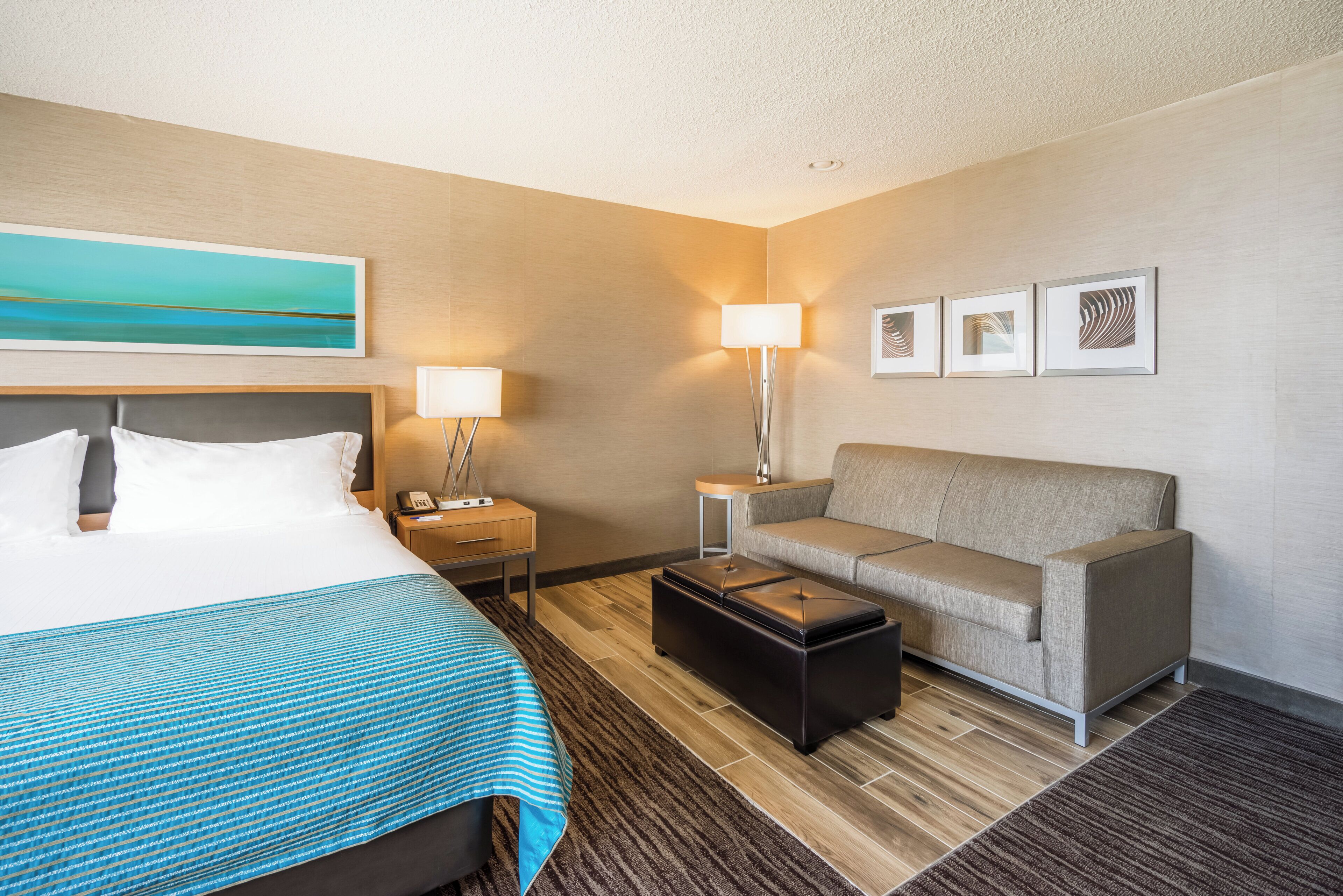 A hotel room with a bed, a sofa, a coffee table, a desk with a lamp, and artwork on the wall.