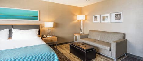 A hotel room with a bed, a sofa, a coffee table, a desk with a lamp, and artwork on the wall.