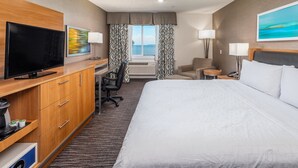 Ein Hotelzimmer mit einem großen Bett, einem Schreibtisch mit Flachbildfernseher, einem Sessel, einem kleinen Tisch mit Lampe und einem Vorhangfenster.