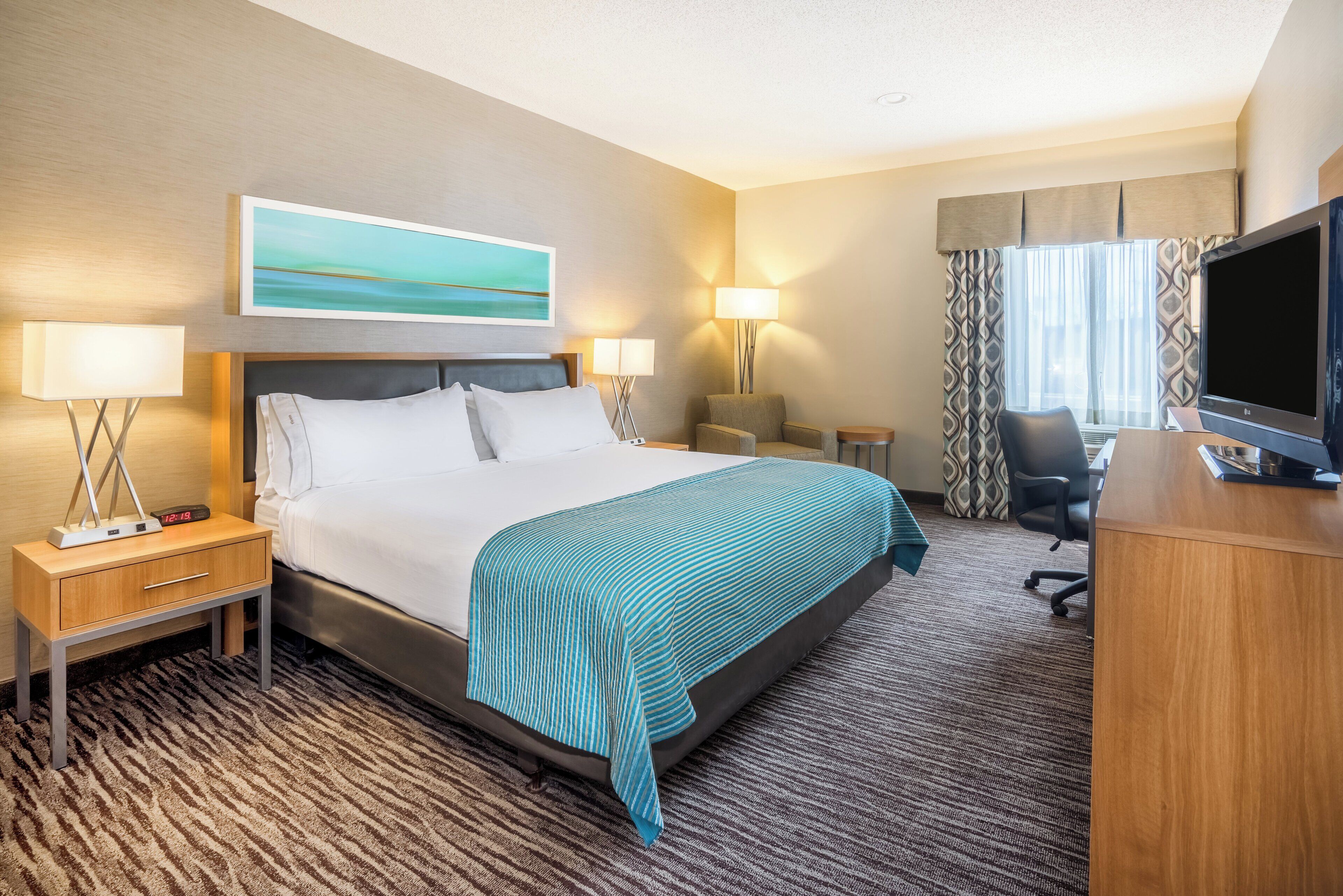 A hotel room with a bed, bedside tables, a desk, a chair, a TV, and a painting on the wall.