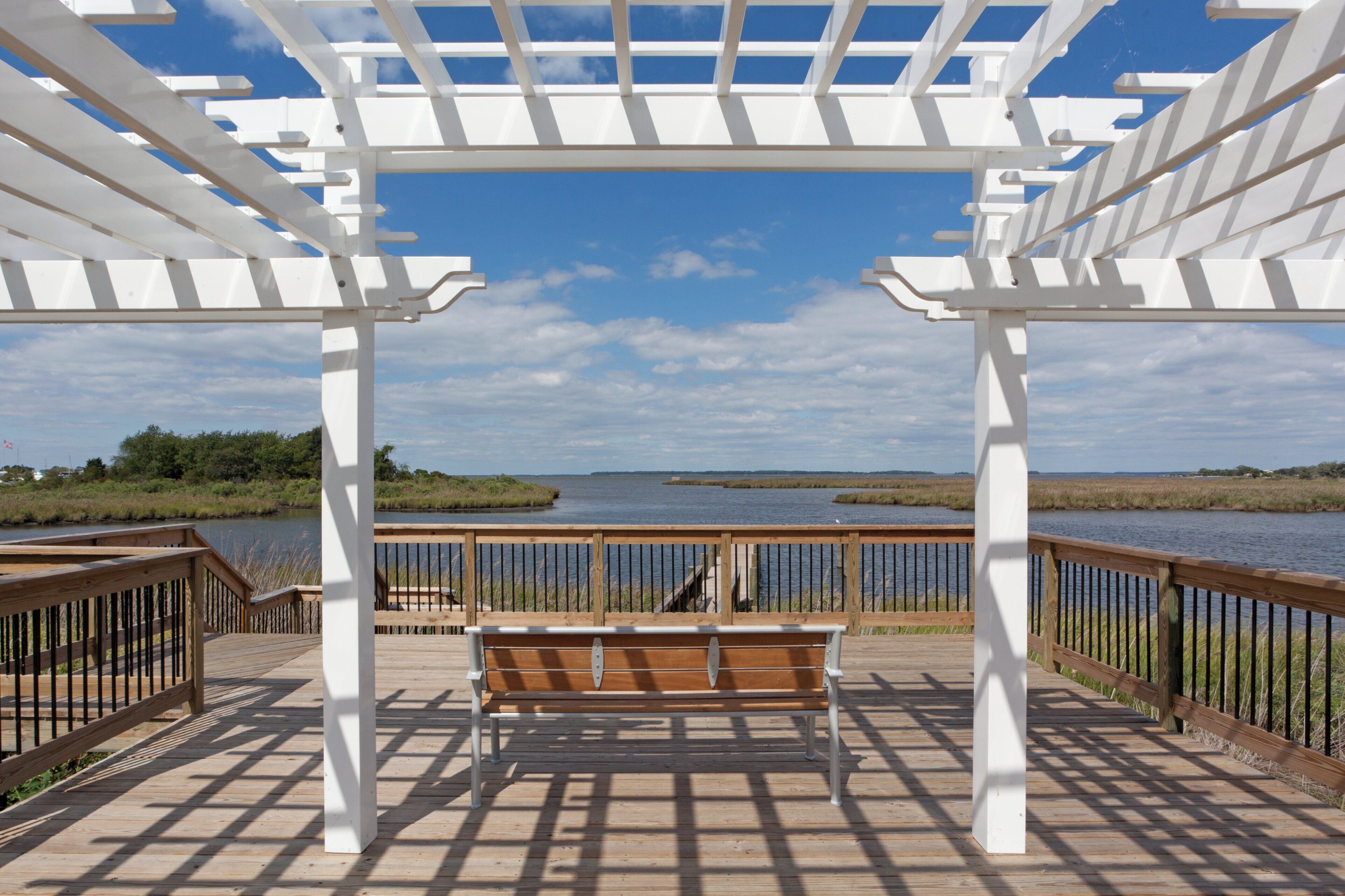Una terraza de madera con una estructura de pérgola blanca, un banco y vistas a un cuerpo de agua.