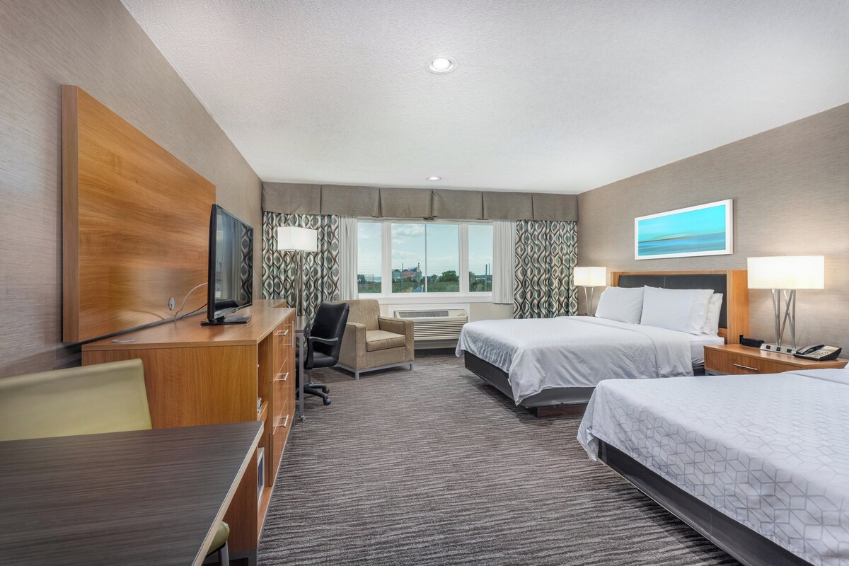 A hotel room with two beds, a desk with a TV, a chair, a desk lamp, and a window with a view.