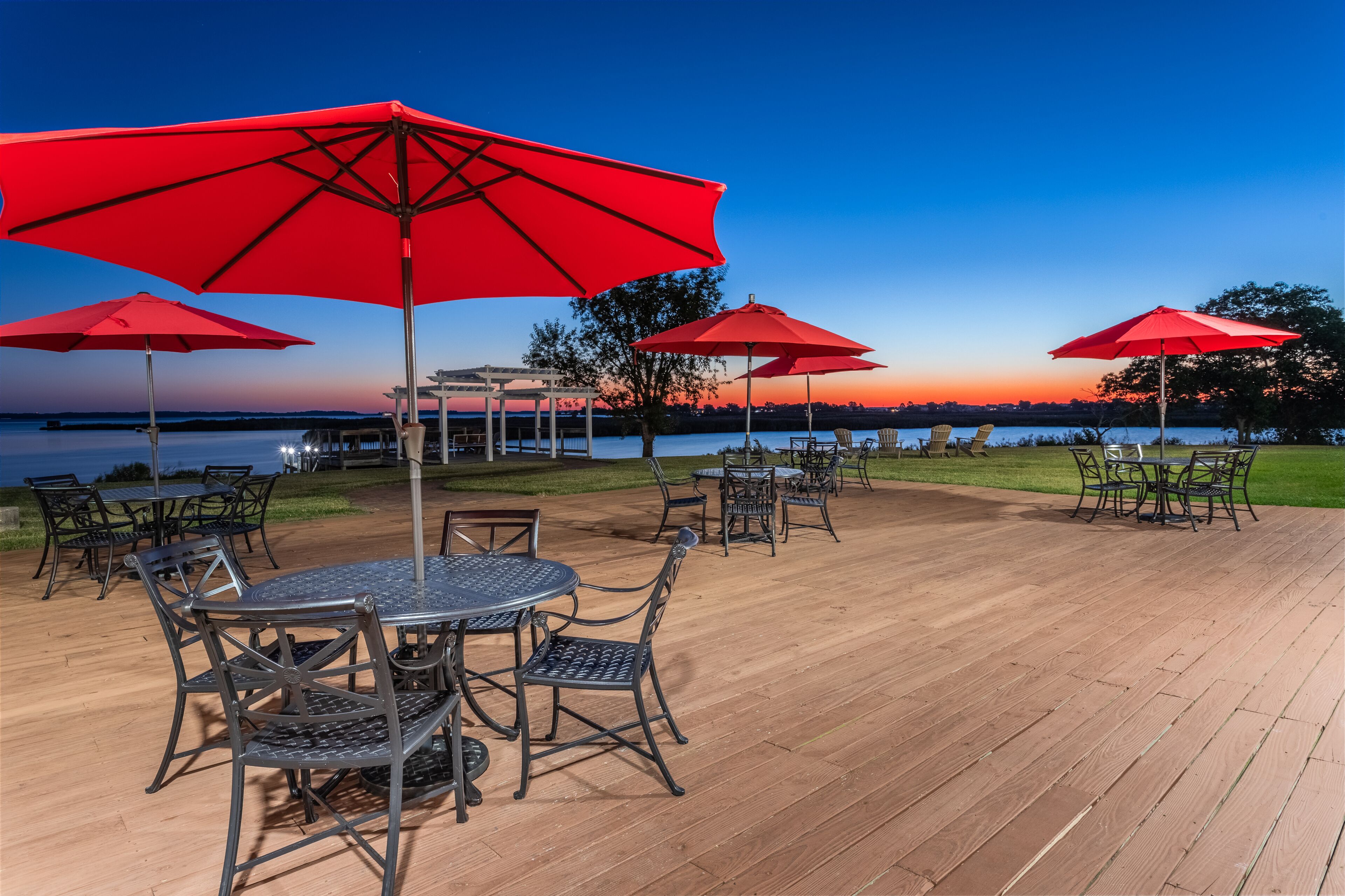 A wooden deck with outdoor seating and red umbrellas, overlooking a body of water.