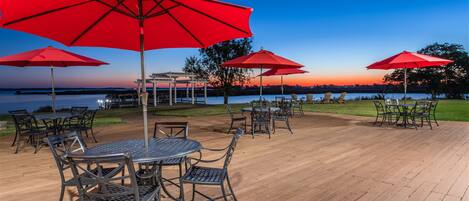 A wooden deck with outdoor seating and red umbrellas, overlooking a body of water.