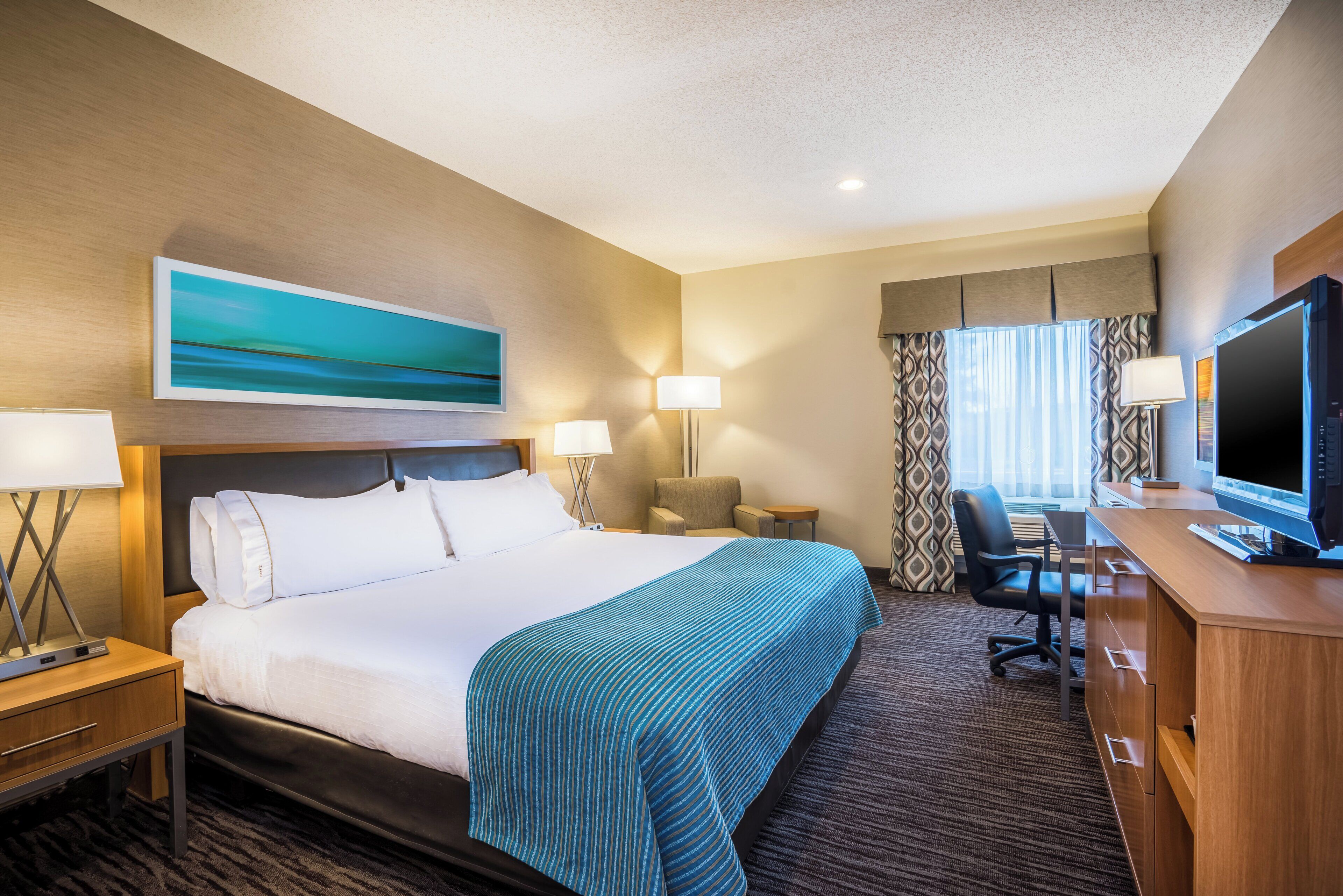 A hotel room with a large bed, two bedside tables, a desk with a computer, a chair, a television, and a window with curtains.
