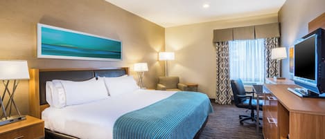 Una habitación de hotel con una cama grande, dos mesitas de noche, un escritorio con computadora, una silla, un televisor y una ventana con cortinas.