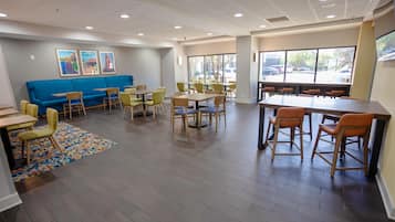 A hotel lobby with a blue sofa, multiple tables and chairs, and large windows offering a view of the parking lot.