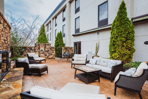 An outdoor patio area with wicker furniture, stone walls, and tall evergreen trees. - Hampton Inn Madison Huntsville Airport (Madison)