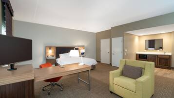 A hotel room with a desk, a bed, a chair, a TV, and a kitchenette.