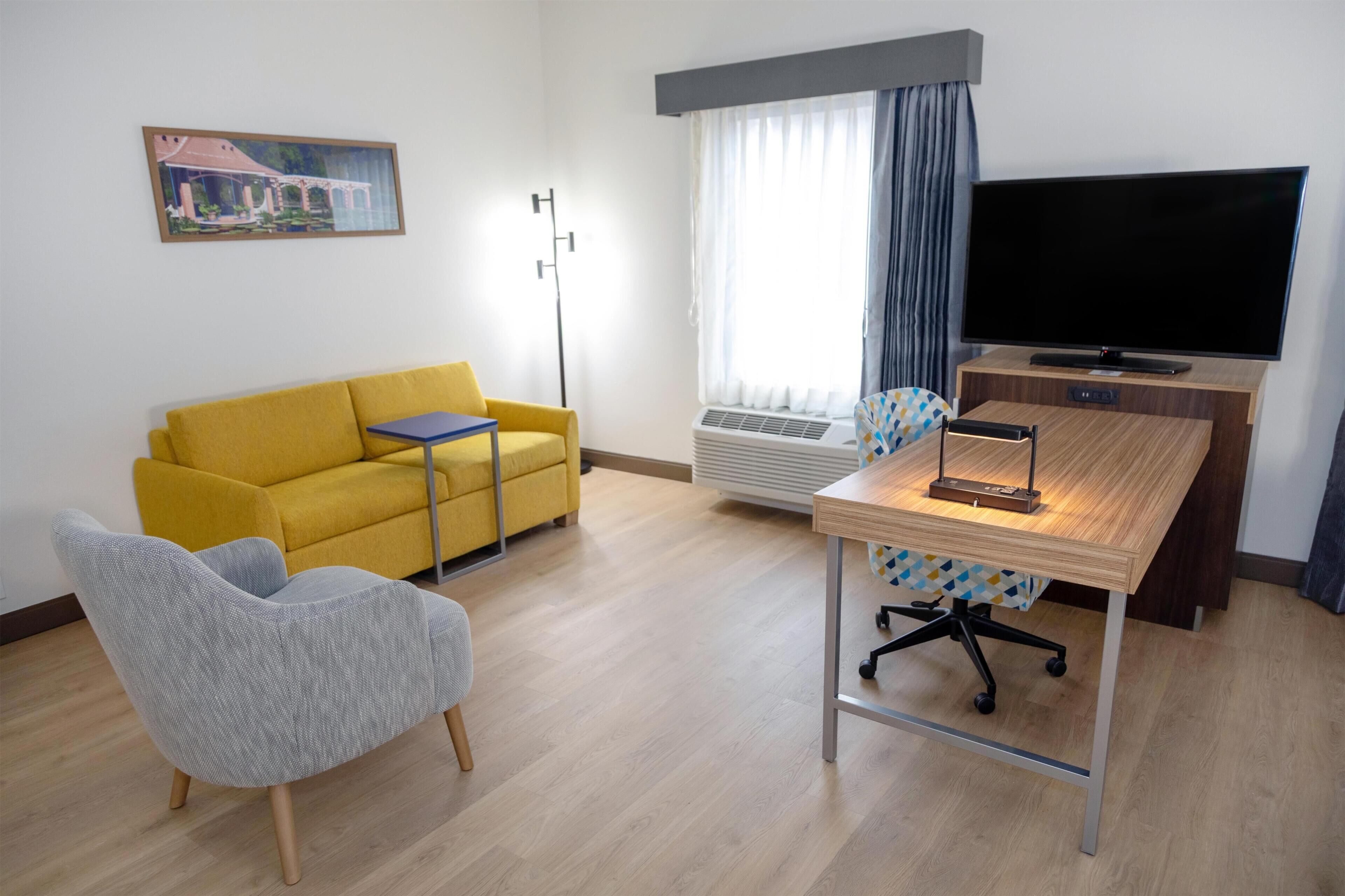 Una habitación con un sofá amarillo, un sillón gris, un escritorio de madera y un televisor de pantalla plana, además de una ventana con cortinas.