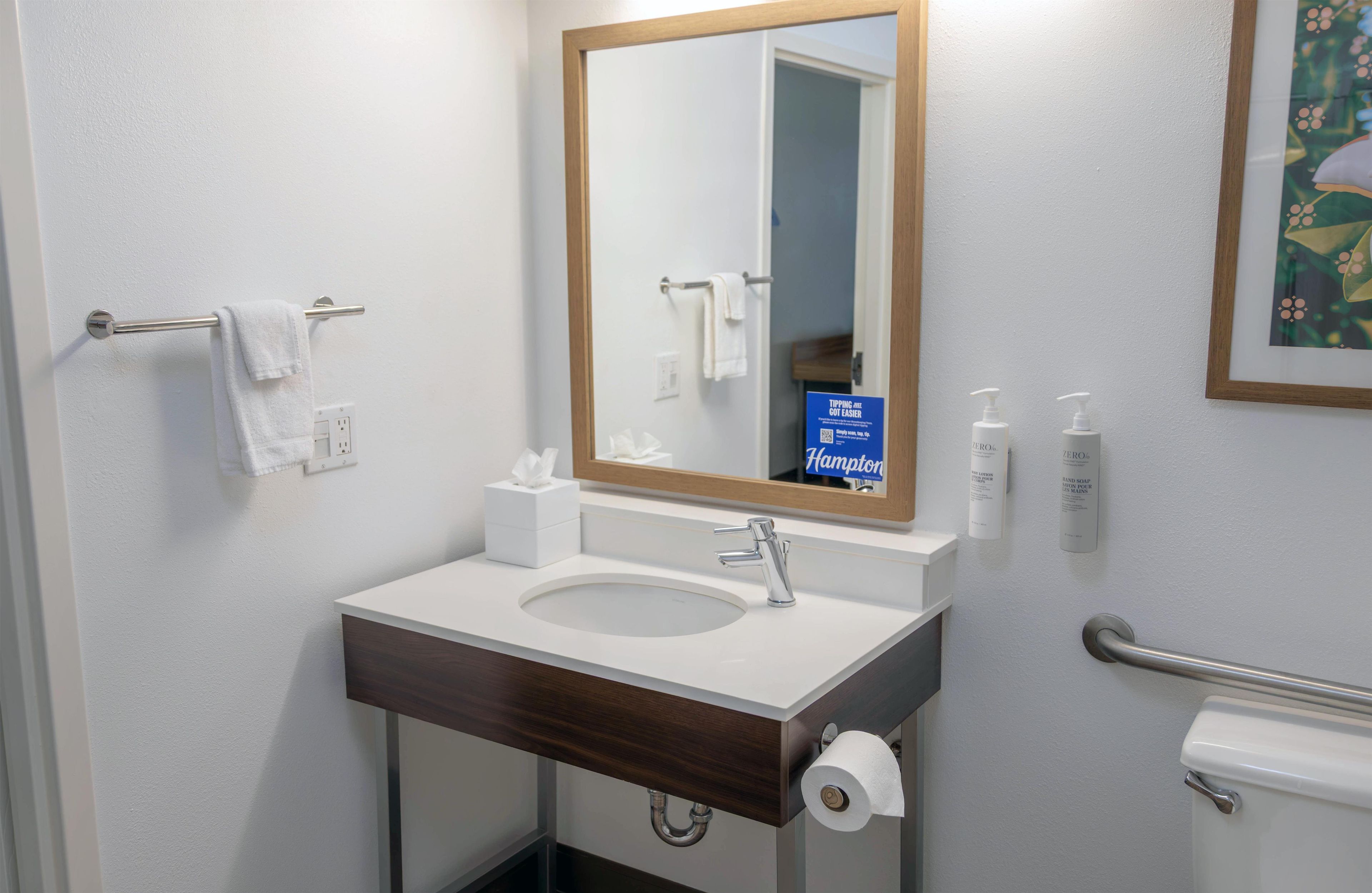 A bathroom with a sink, mirror, towel rack, toilet paper, and hand sanitizer.