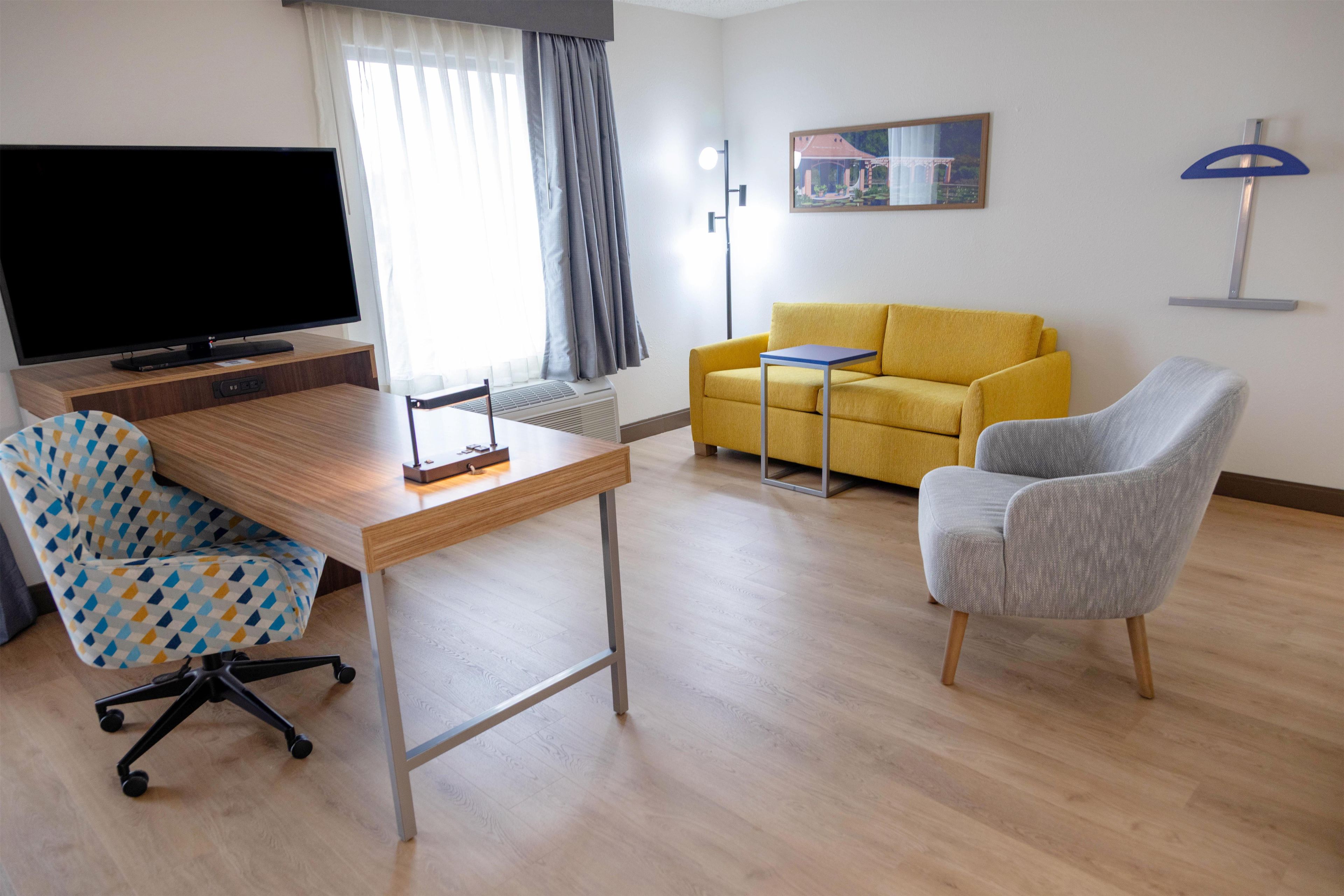 Une chambre d’hôtel moderne équipée d’un téléviseur à écran plat, d’un bureau en bois, d’un canapé jaune, d’un fauteuil gris et d’un tableau accroché au mur.