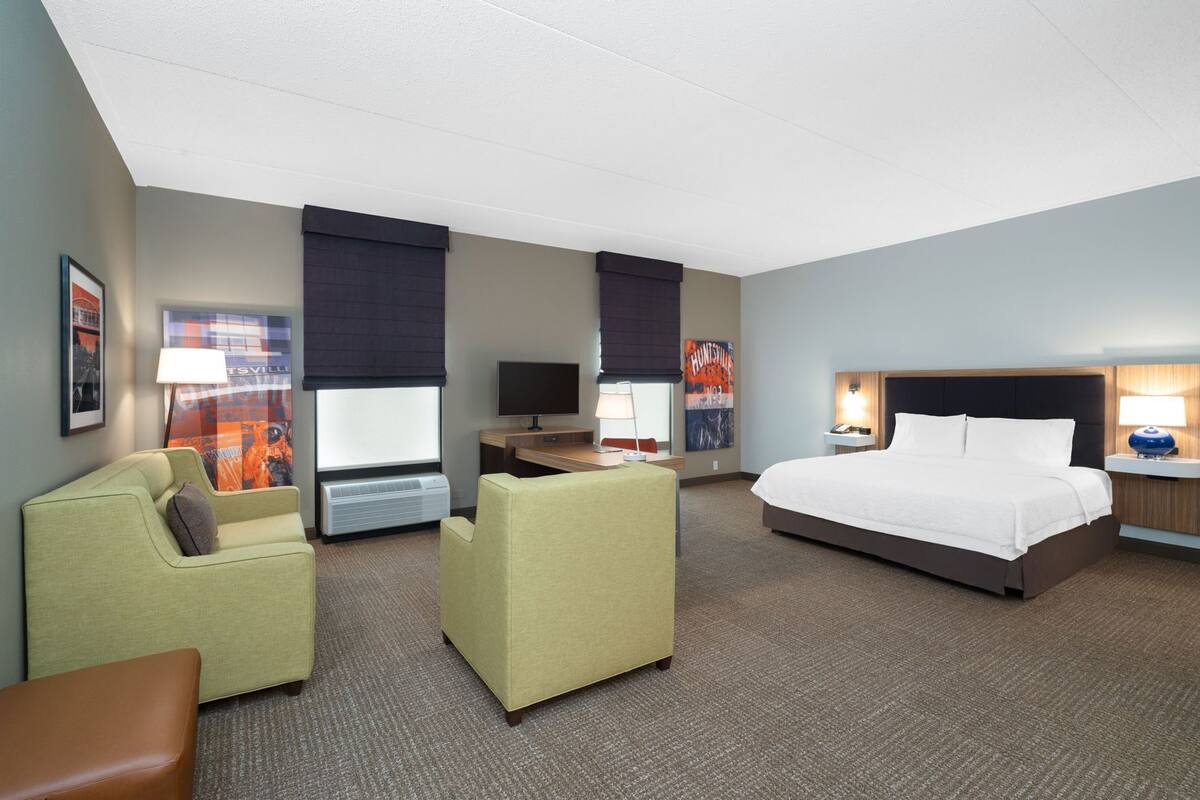 A hotel room with a bed, two armchairs, a desk with a computer, a flat-screen TV, and a window with a view of a cityscape.