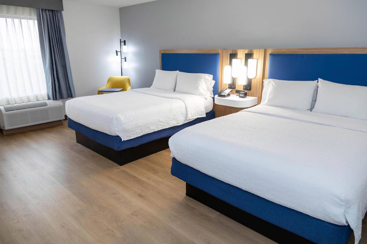 A hotel room with two beds, a bedside table with a lamp, a chair, and a window with curtains.