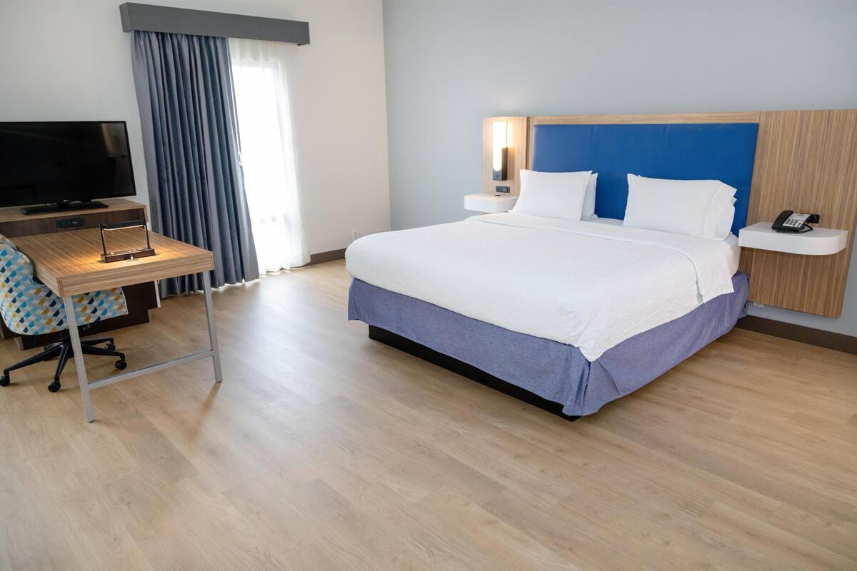 A hotel room with a bed, a desk with a TV, a chair, a phone, and a window with curtains.