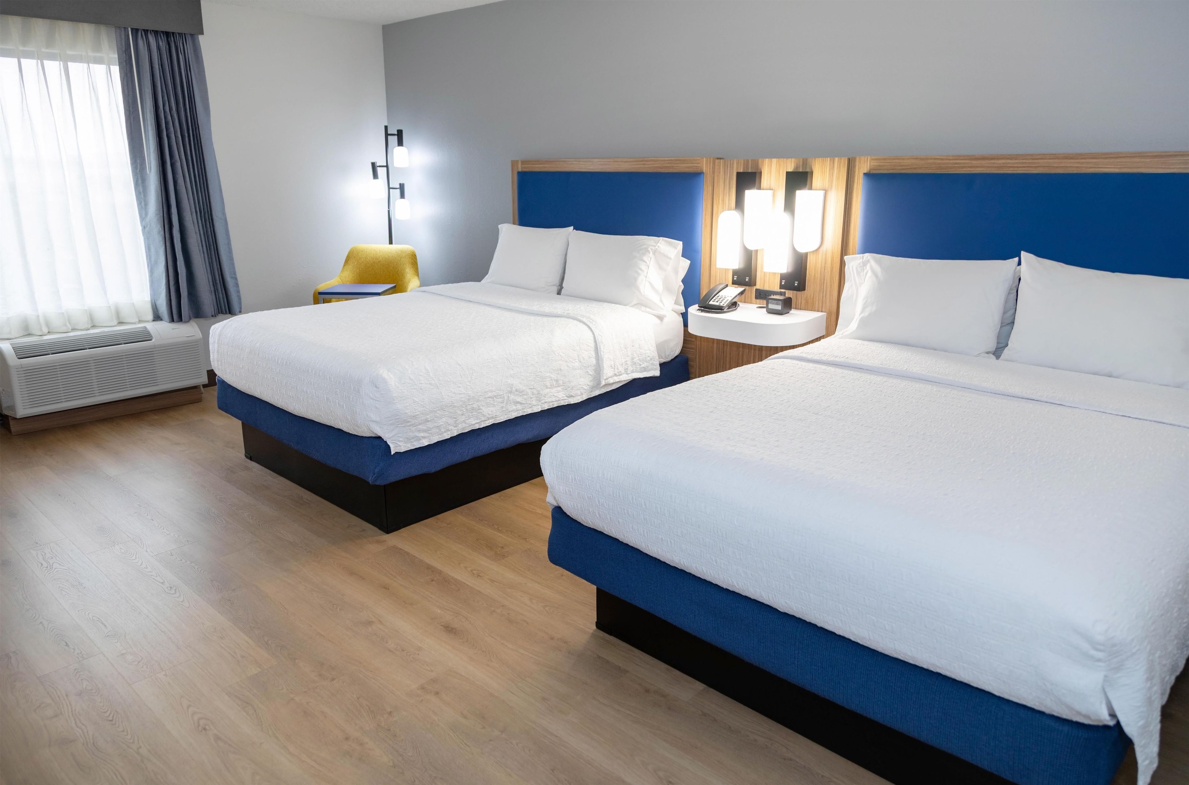 A hotel room with two beds, a bedside table with a lamp, a chair, and a window with curtains.
