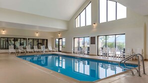 An indoor swimming pool with clear glass walls, white lounge chairs, and a high ceiling with skylights.