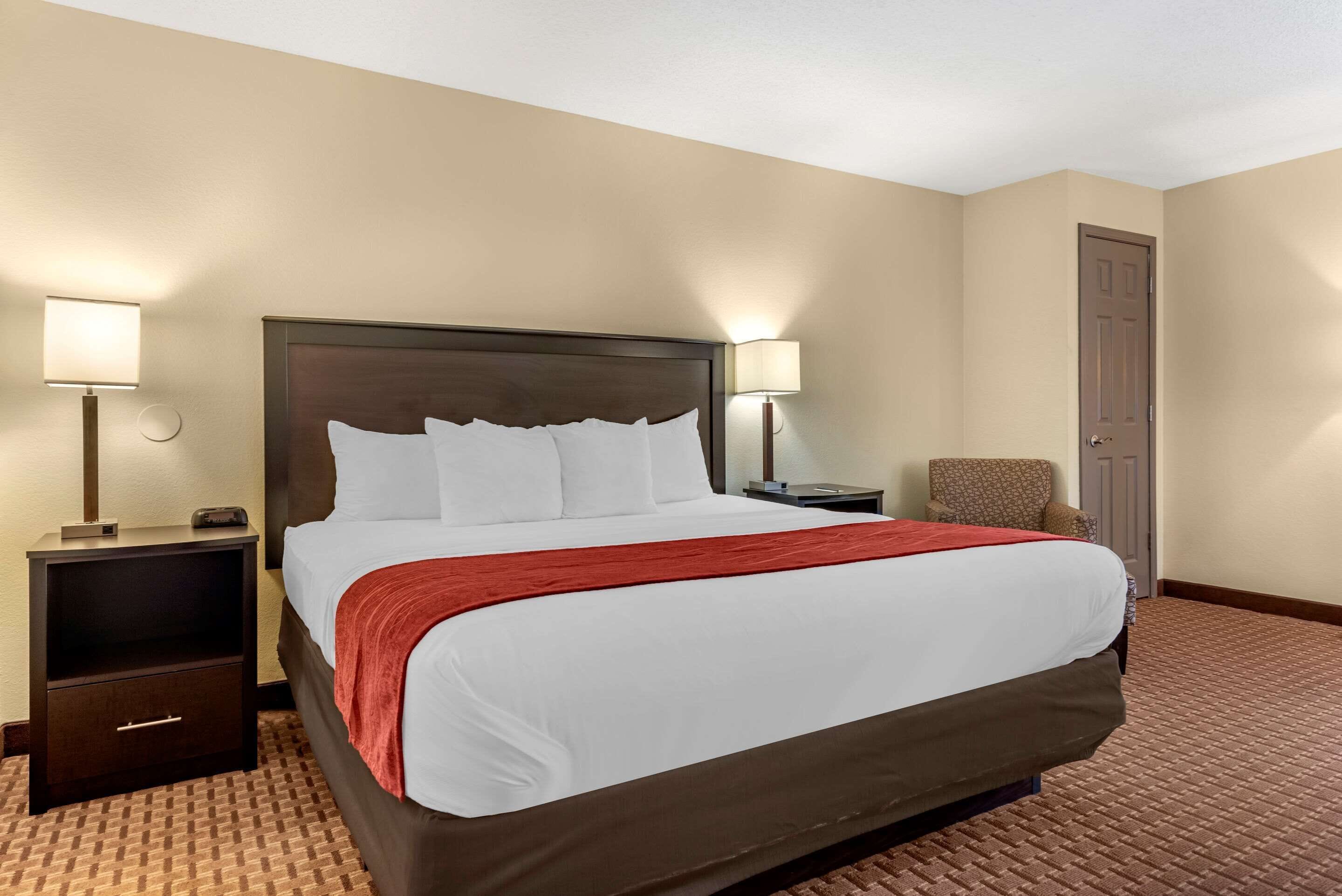 Quarto de hotel com uma cama grande, duas mesas de cabeceira com abajures, uma cadeira e uma porta.