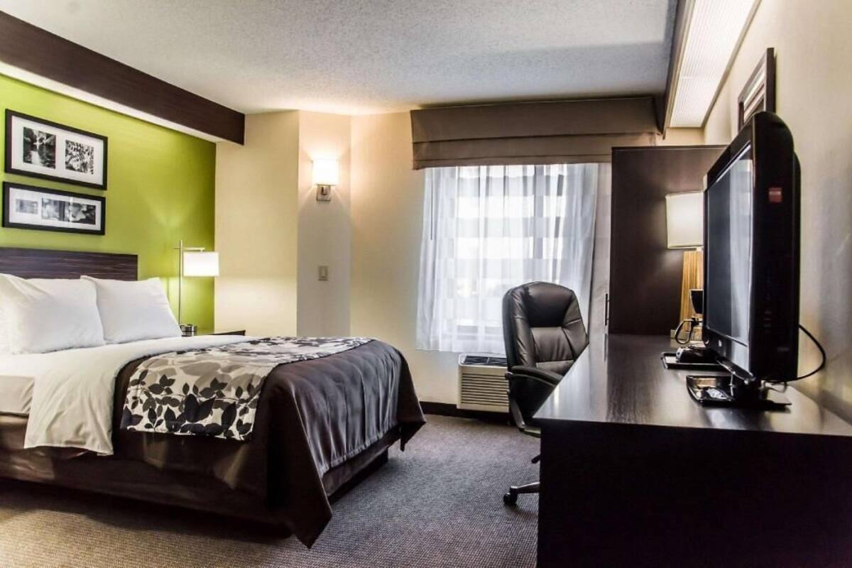 Ein Hotelzimmer mit einem Bett, einem Schreibtisch mit Fernseher, einem Sessel und einem Fenster mit Vorhängen.