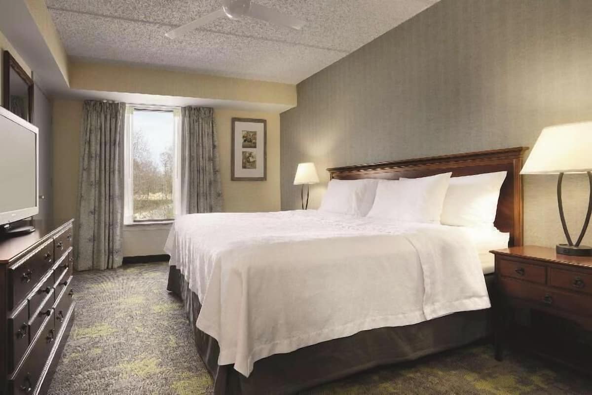 Ein Hotelzimmer mit einem Bett, Nachttischlampen, einem Schminktisch, einem Fernseher, einem Fenster mit Vorhängen und einem gerahmten Bild an der Wand.