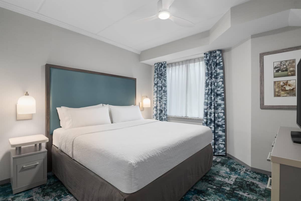 Camera d'albergo con un letto grande, un comodino, una scrivania, una ventola a soffitto e una finestra con tende.