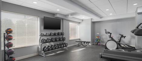 Ein Fitnessraum mit Trainingsgeräten, einem Fernseher und einem Fenster mit Jalousien.