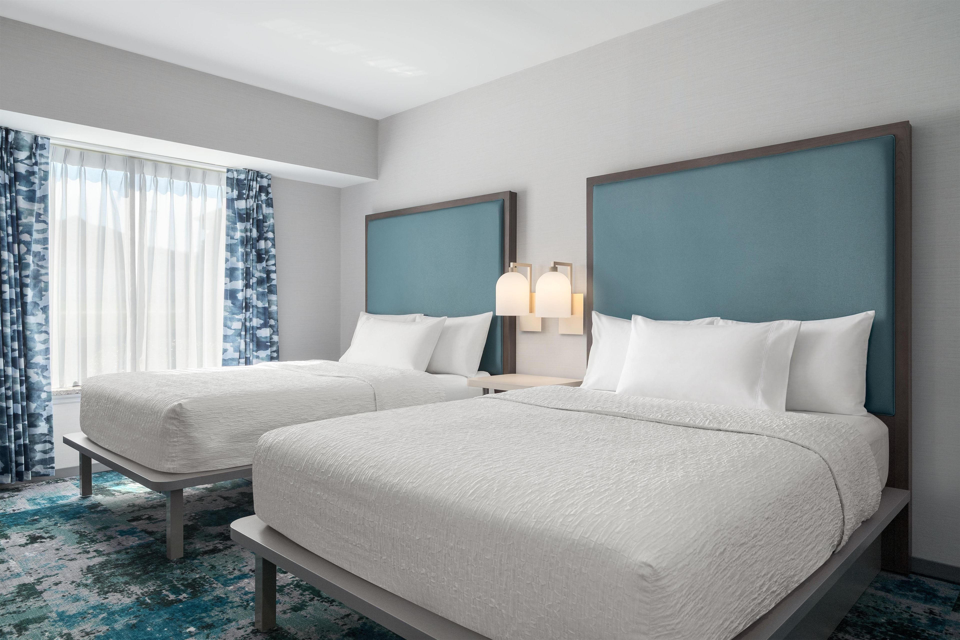 Habitación de hotel con dos camas, una mesita de noche, una lámpara y una ventana con cortinas.