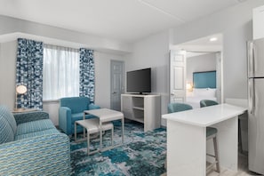 A compact living space with a sofa, a small TV, a desk, and a bed with a headboard. - Homewood Suites by Hilton Williamsburg (Williamsburg)