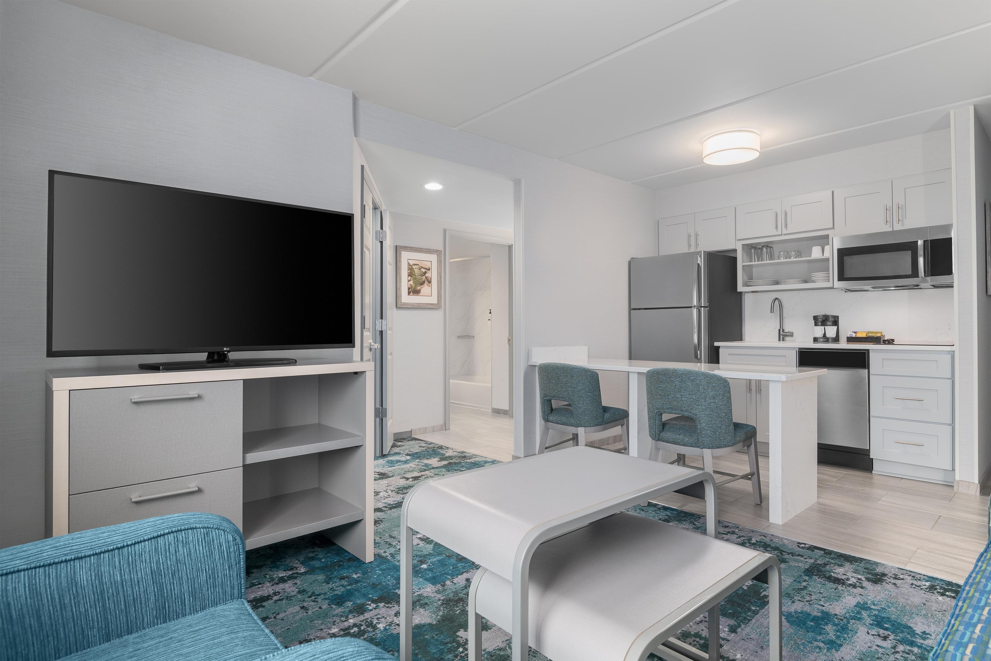 A modern hotel room with a flat-screen TV, a dining area with two chairs, a kitchen with a microwave, refrigerator, and sink.