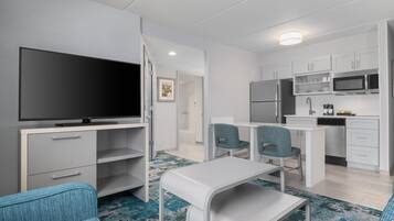 A modern hotel room with a flat-screen TV, a dining area with two chairs, a kitchen with a microwave, refrigerator, and sink.