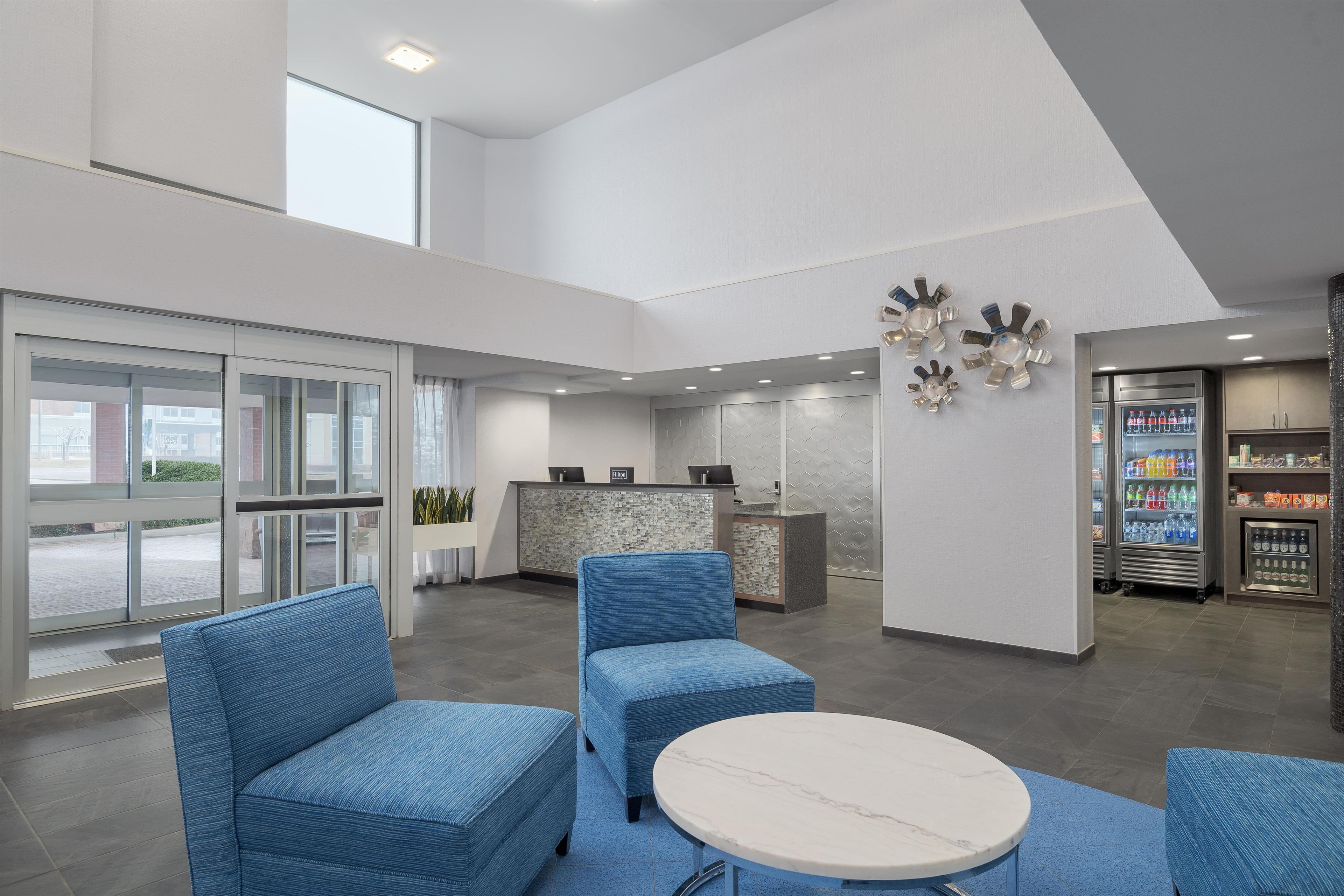 A modern hotel lobby with seating, a reception desk, and a minibar.