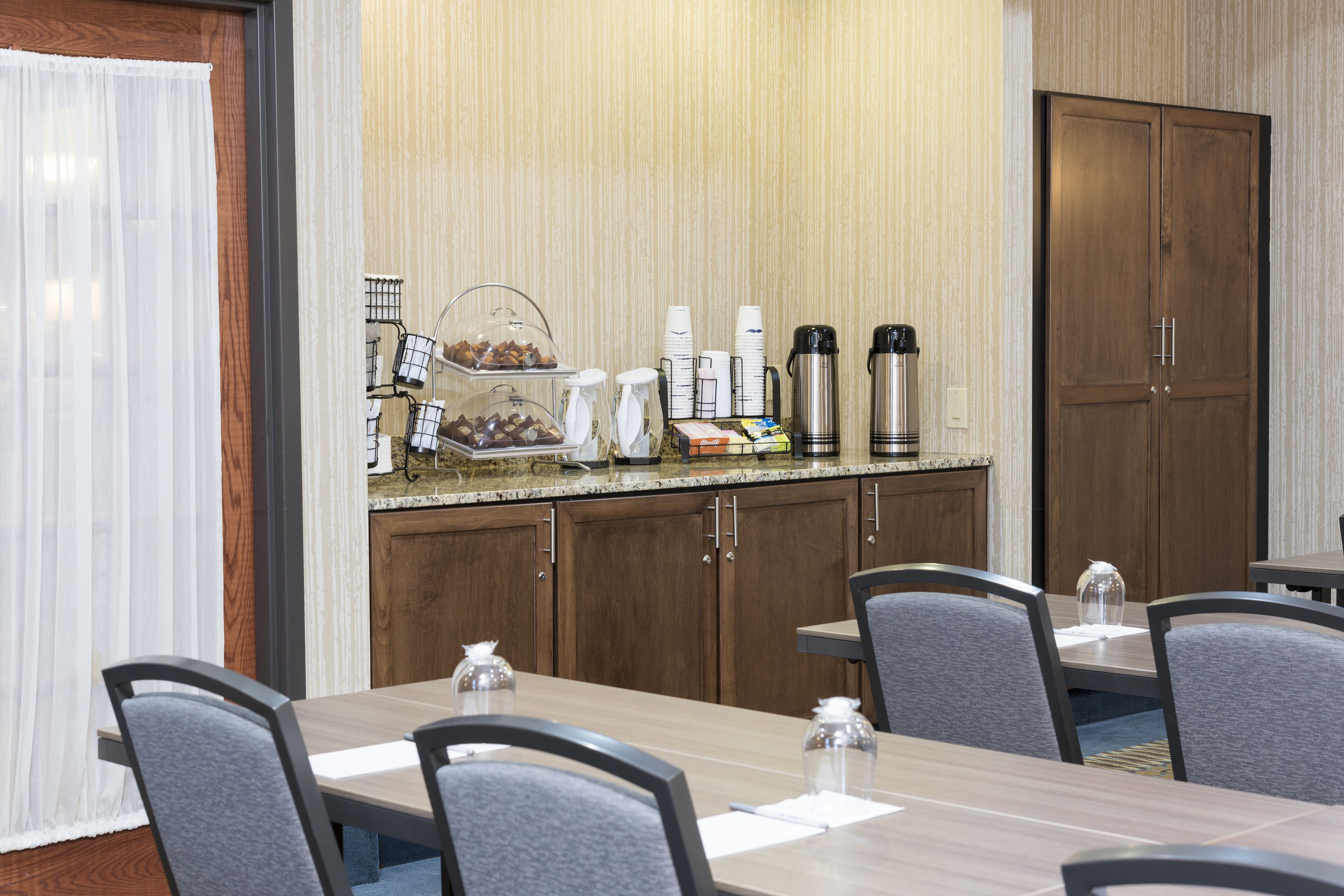A hotel dining area with a long table, chairs, and a breakfast bar with coffee makers and condiments.