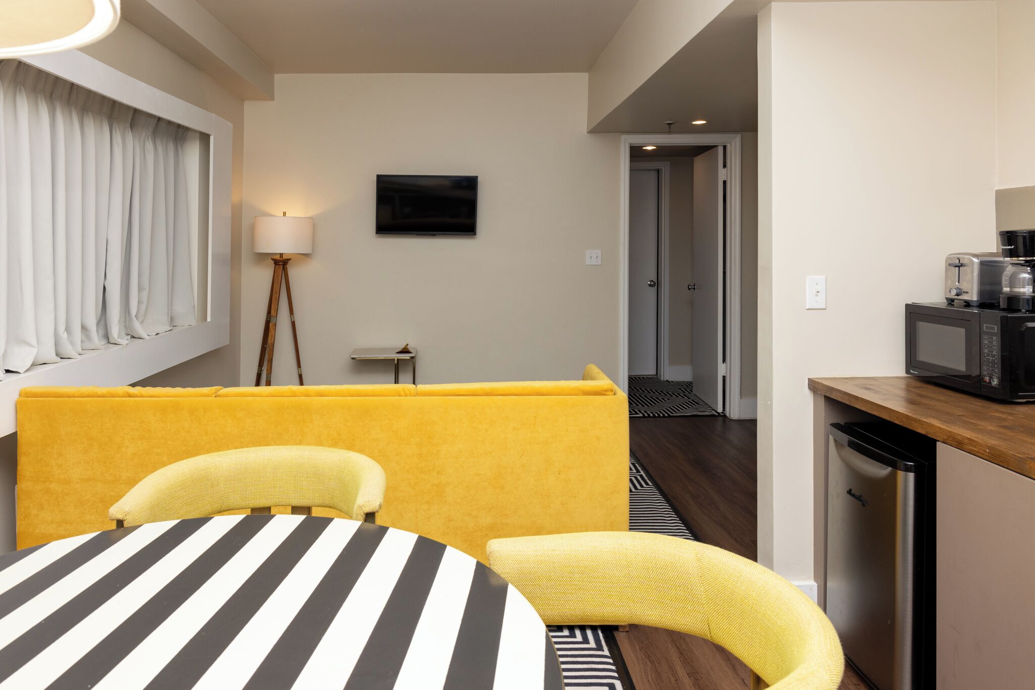 Quarto de hotel moderno com sofá amarelo, mesa listrada e cozinha equipada com micro-ondas e cafeteira.