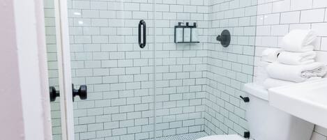 Un baño moderno con paredes de azulejos blancos, cerramiento de ducha de vidrio, inodoro blanco y lavabo con una pila de toallas.