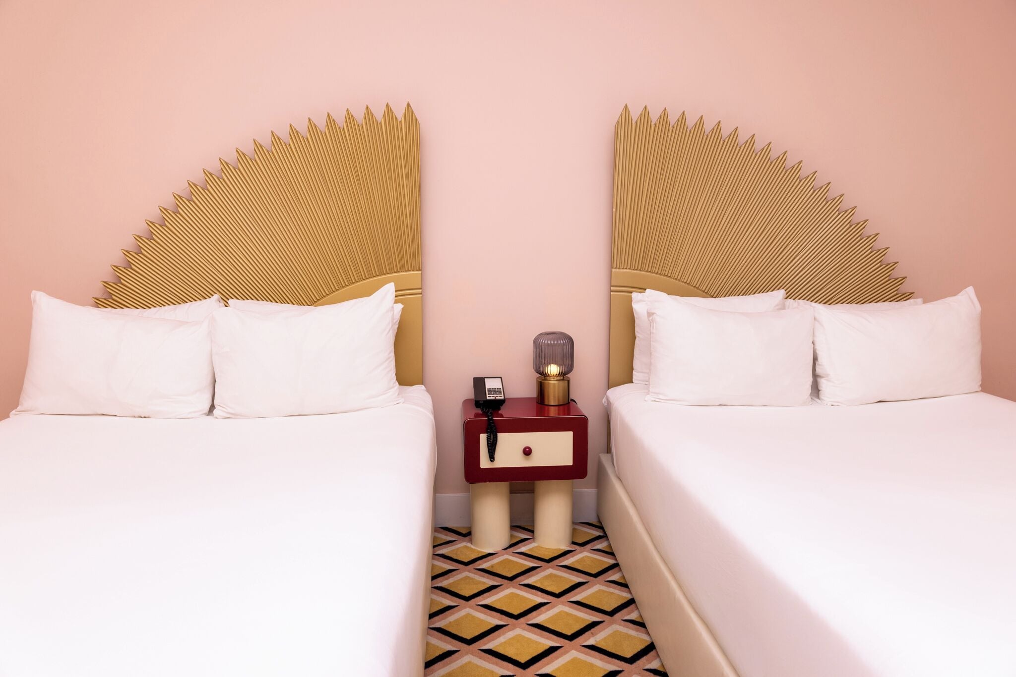 A hotel room with two beds, each with a unique headboard design, a bedside table with a lamp, and a patterned floor.