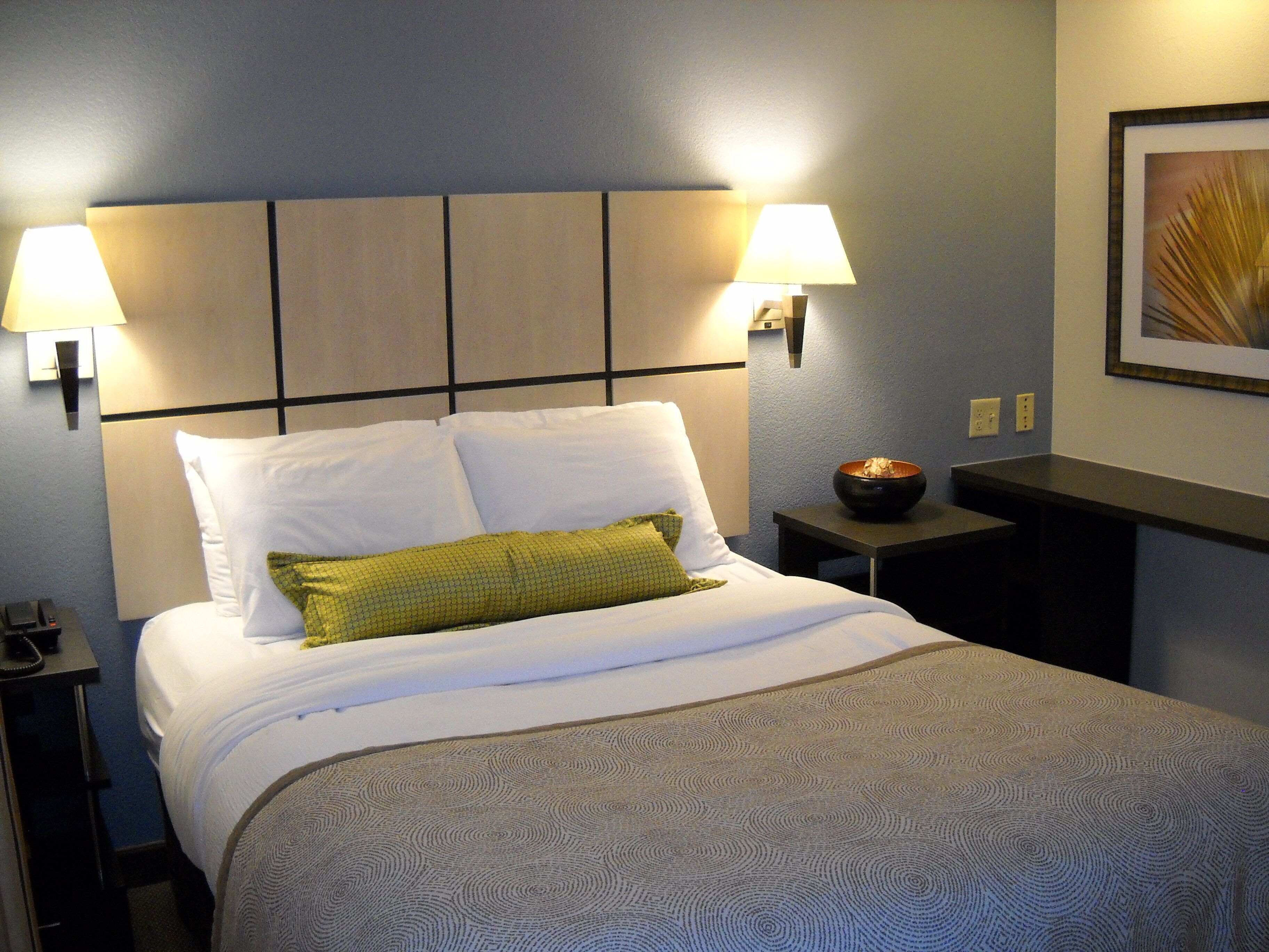Habitación de hotel con una cama, dos lámparas de noche, una mesita de noche con un cuenco y un cuadro en la pared.