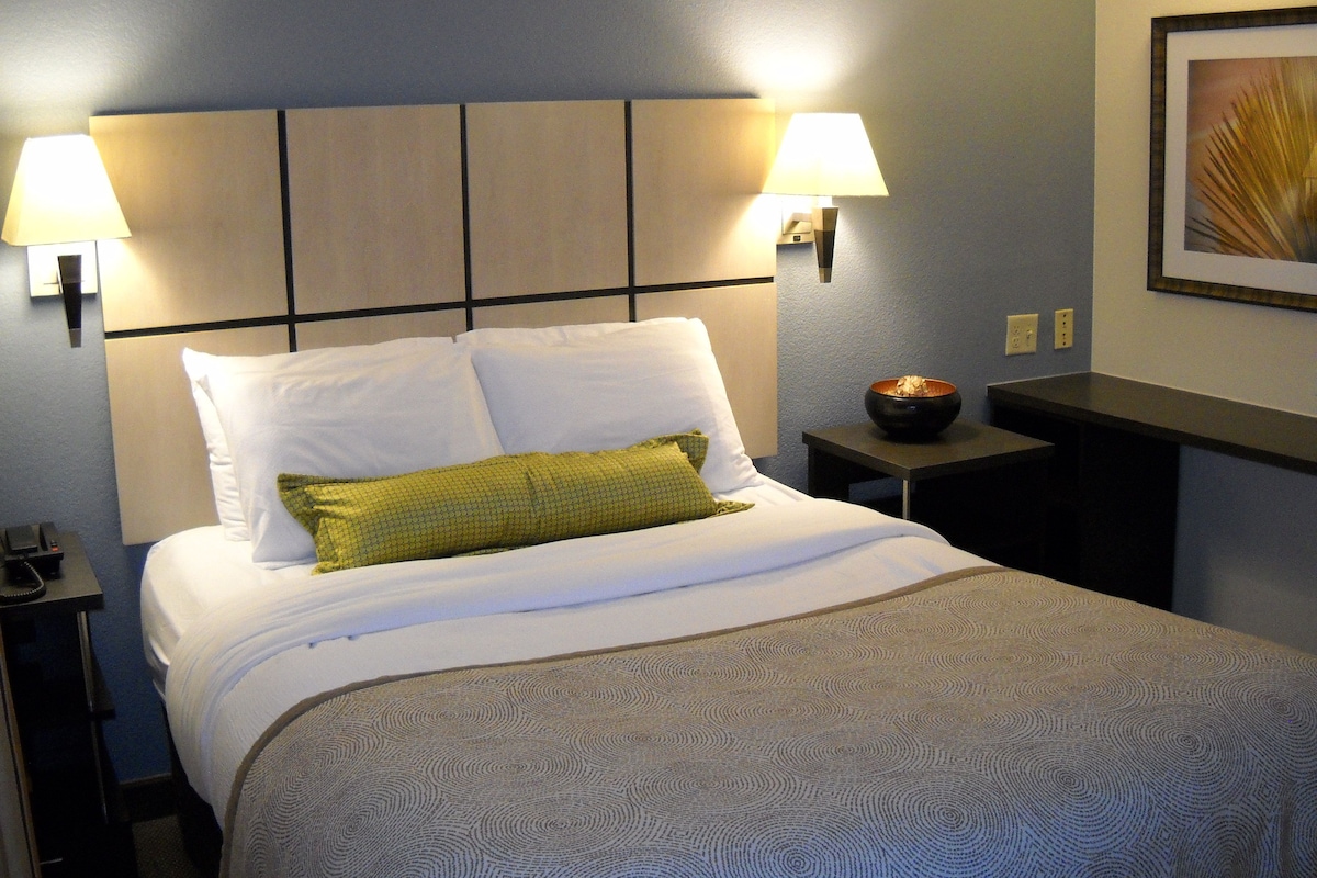 Habitación de hotel con una cama, dos lámparas de noche, una mesita de noche con un cuenco y un cuadro en la pared.